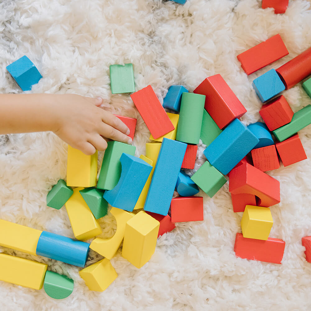 Melissa and Doug 100 Piece Wood Blocks Set pieces on a rug with a little childs hand on the pieces